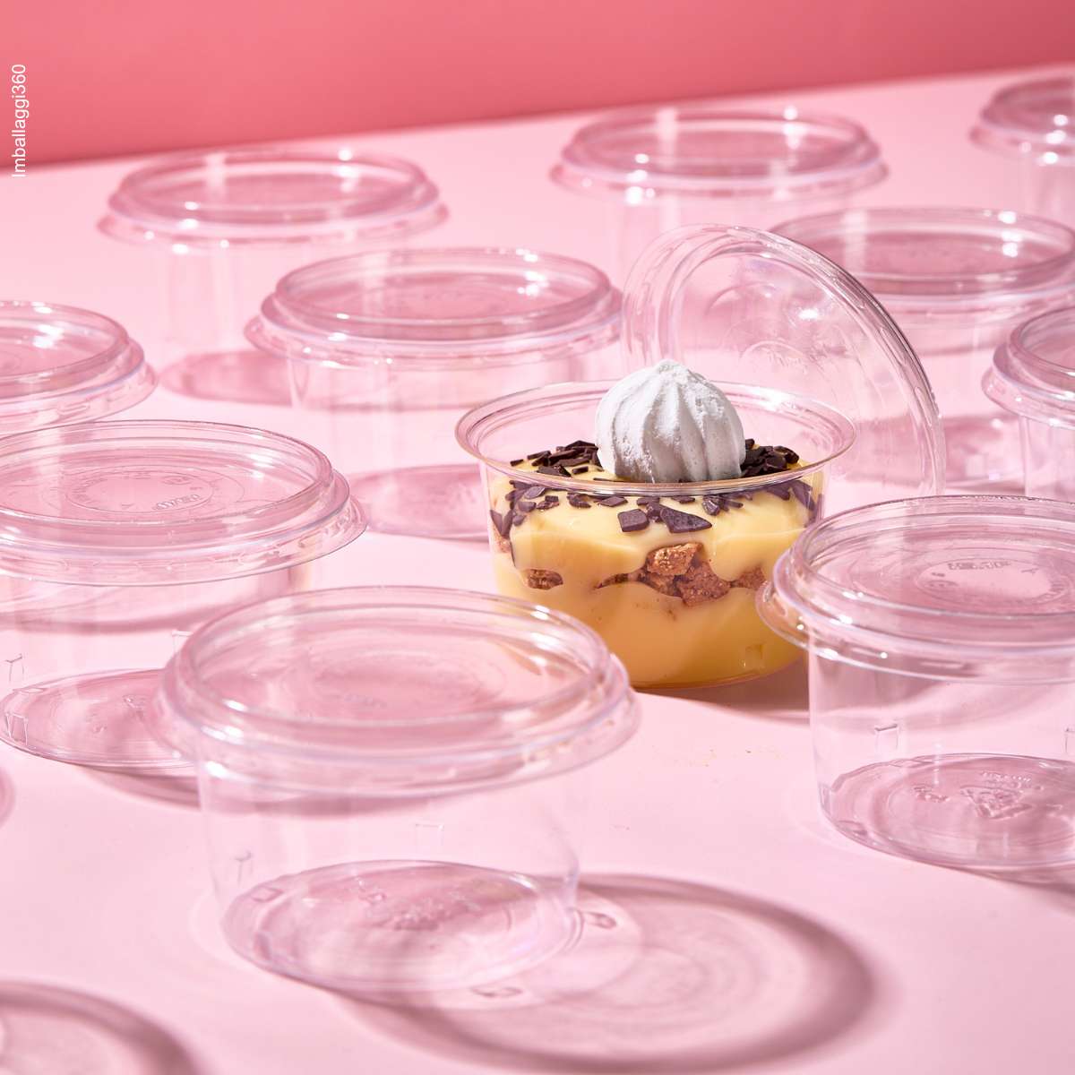 Coppetta per monoporzioni, piena di dolce e aperta, tra tante coppette monoporzione chiuse e vuote, su sfondo rosa