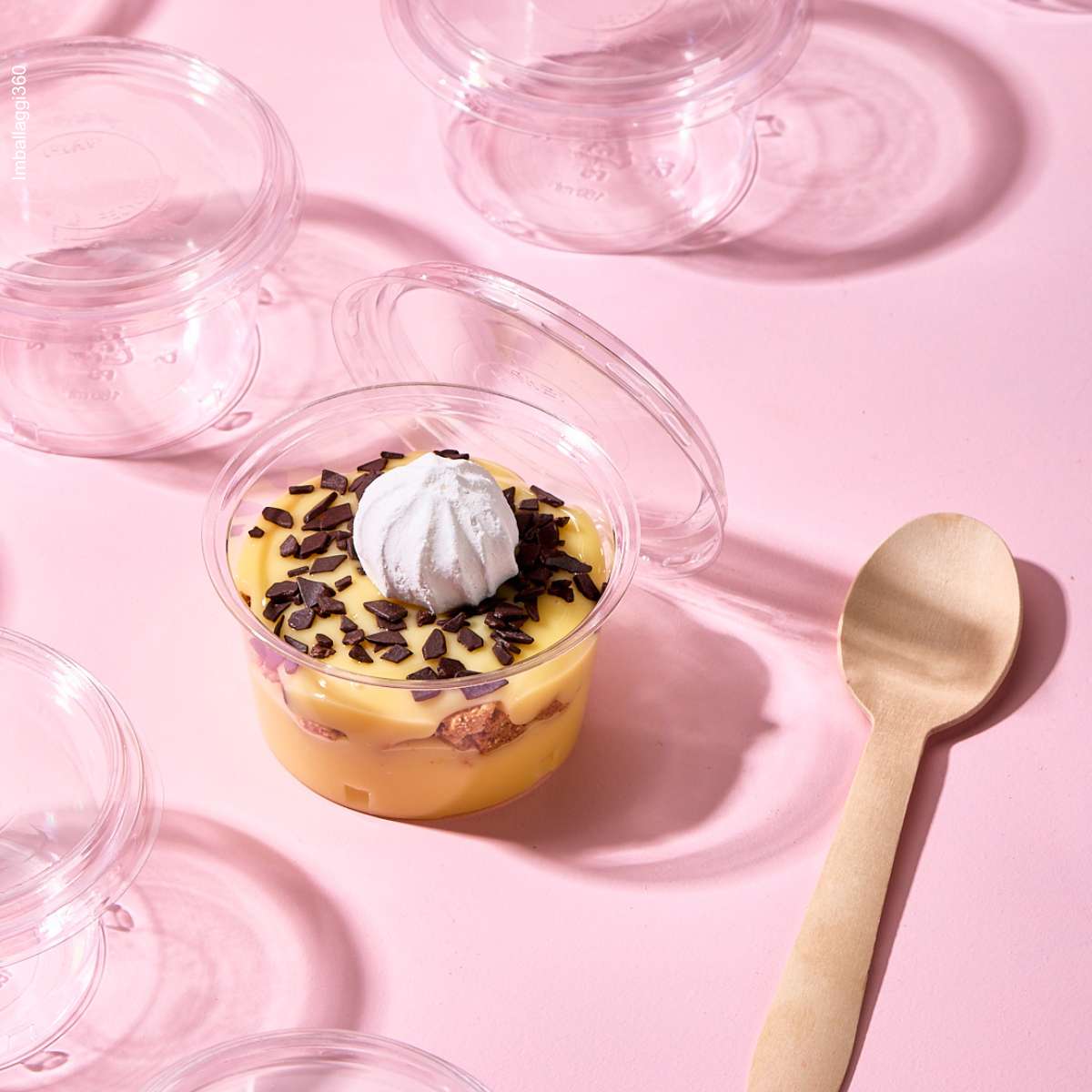 Coppetta per monoporzioni, piena di dolce e aperta, vicino ad un cucchiaio di legno tra tante coppette monoporzione chiuse e vuote, su sfondo rosa