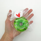 Mano che tiene nel palmo busta trasparente chiusa con etichetta tonda personalizzata con all'interno una ciambella verde 