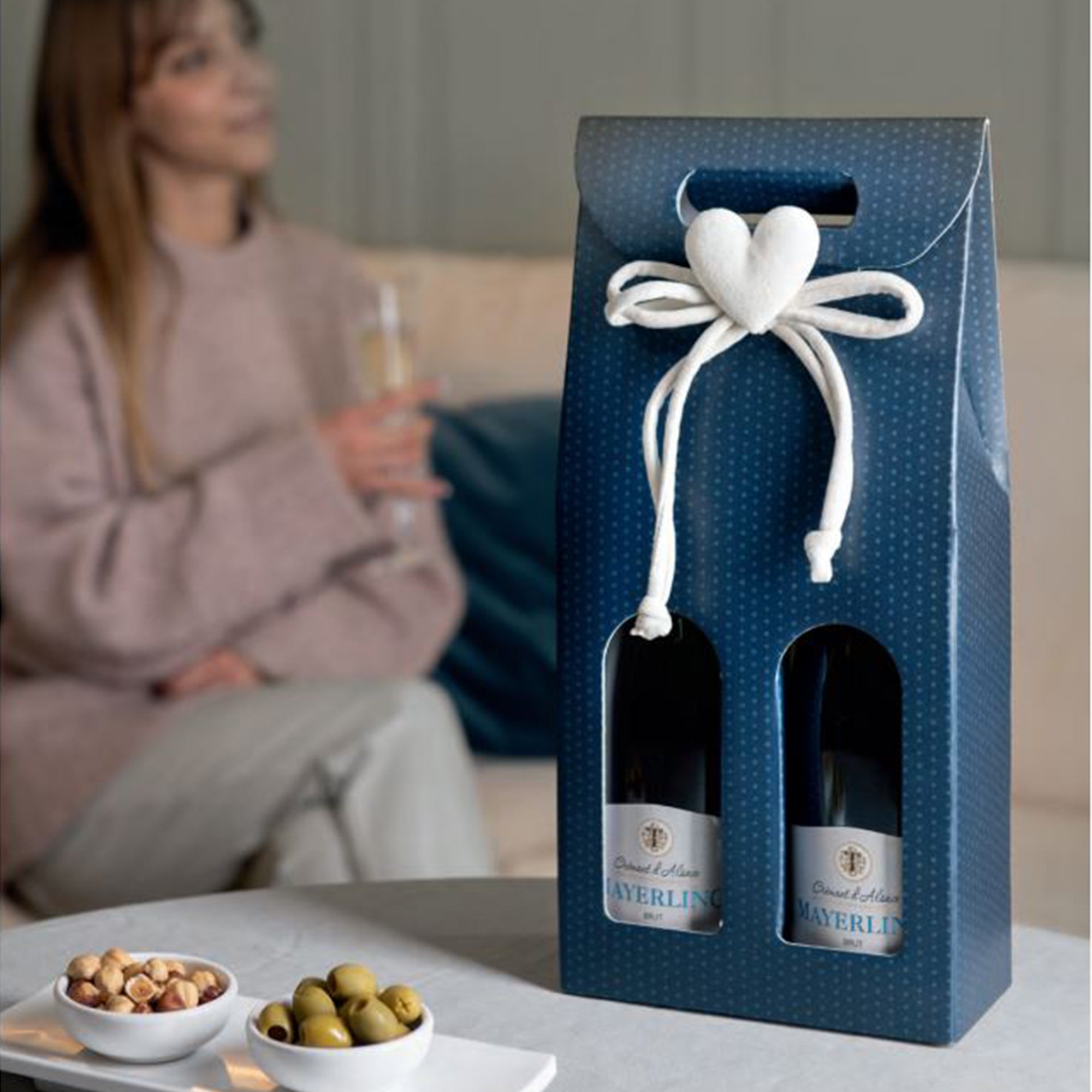 Confezione di cartone per due vini, con finestra, fantasia blu a pois bianchi, su un tavolino mentre una ragazza tiene tra e mani un bicchiere di vino bianco sullo sfondo