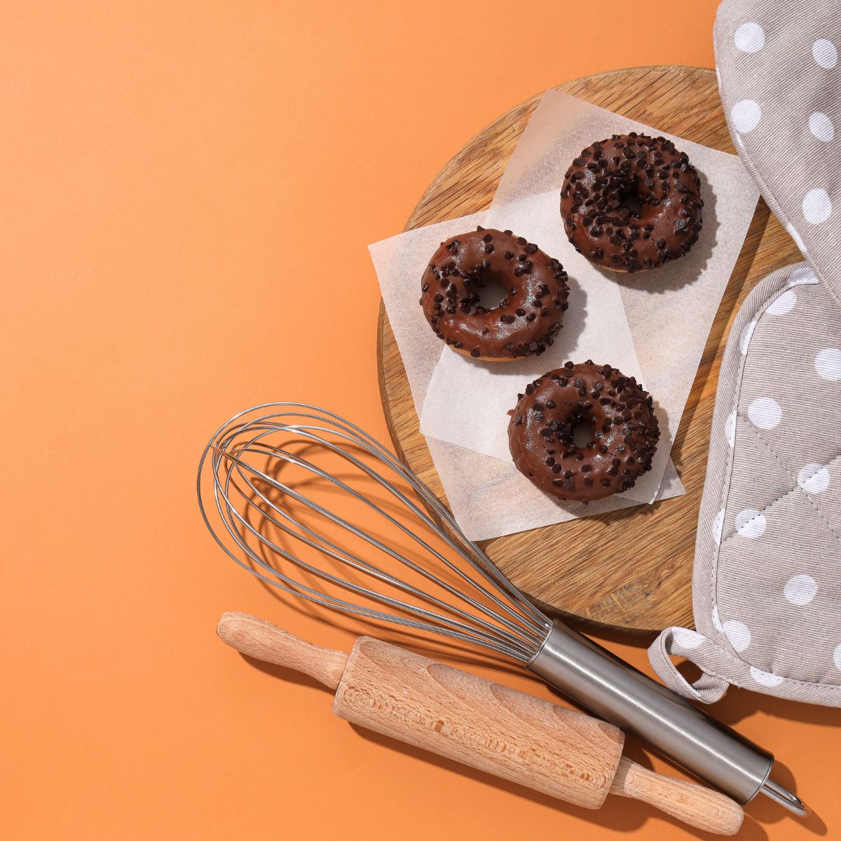 Elementi da cucina: frusta, mattarello, tagliere con carta antigrasso e donuts, presine. Tutto su sfondo arancione