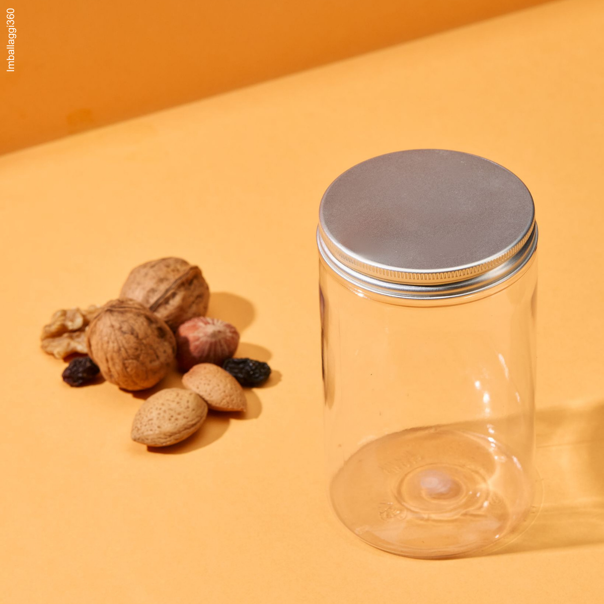 Barattolo trasparente con tappo di alluminio, chiuso. vuoto, accanto a frutta secca su sfondo arancione