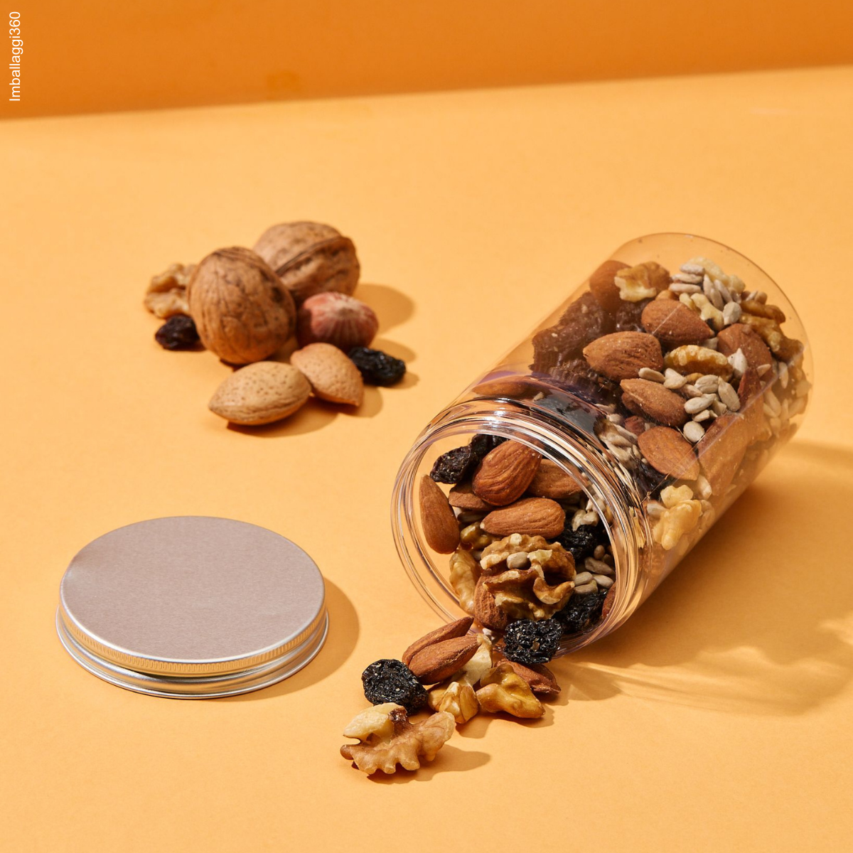 Barattolo trasparente con tappo di alluminio, aperti e pieno di frutta secca. Rovesciato. tappo vicino accanto a frutta secca su sfondo arancione. 