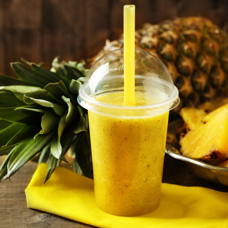 Bicchiere di plastica con coperchio a cupola e cannuccia gialla, pieno di succo di ananas, vicino ad un ananas su un tovagliolo giallo 