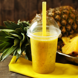 Bicchiere di plastica con coperchio a cupola e cannuccia gialla, pieno di succo di ananas, vicino ad un ananas su un tovagliolo giallo 