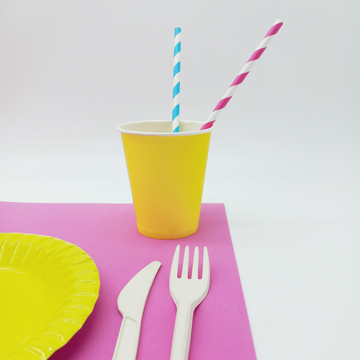 Bicchiere di carta giallo con dentro due cannucce, una bianca e blu ed una rosa e blu, vicino al dettaglio di un piatto di carta giallo, con posate di plastica su tovaglietta americana rosa 