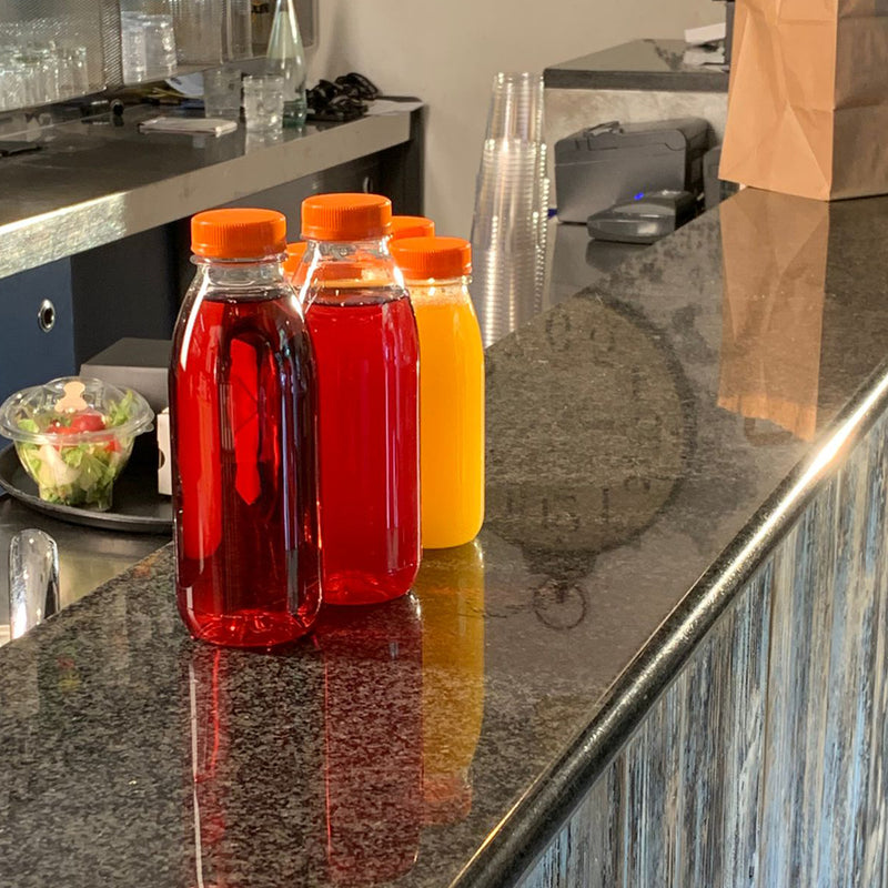 Bottigliette per succo, ripiene di vari succhi. Tappo arancione. Disposte sul bacone di un bar