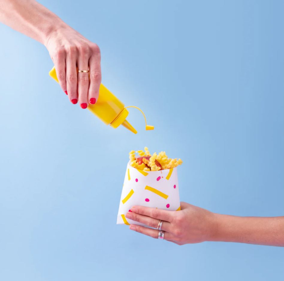 Sacchetto porta patatine retto da una mano, pieno di patatine e altra mano che versa maionese
