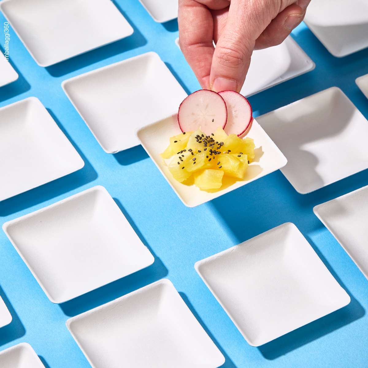 Piattino monoporzione in pola di cellulosa, tenuta alzata tra due dita ripieno di finger food, accanto a piattini vuoti su sfondo azzurro