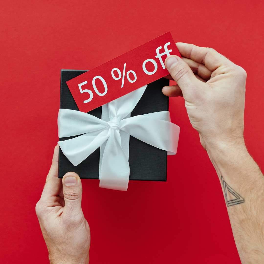 Pacco nero con nastro bianco su sfondo rosso tenuto da due mani che reggono anche un cartellino rosso con su scritto 50% off