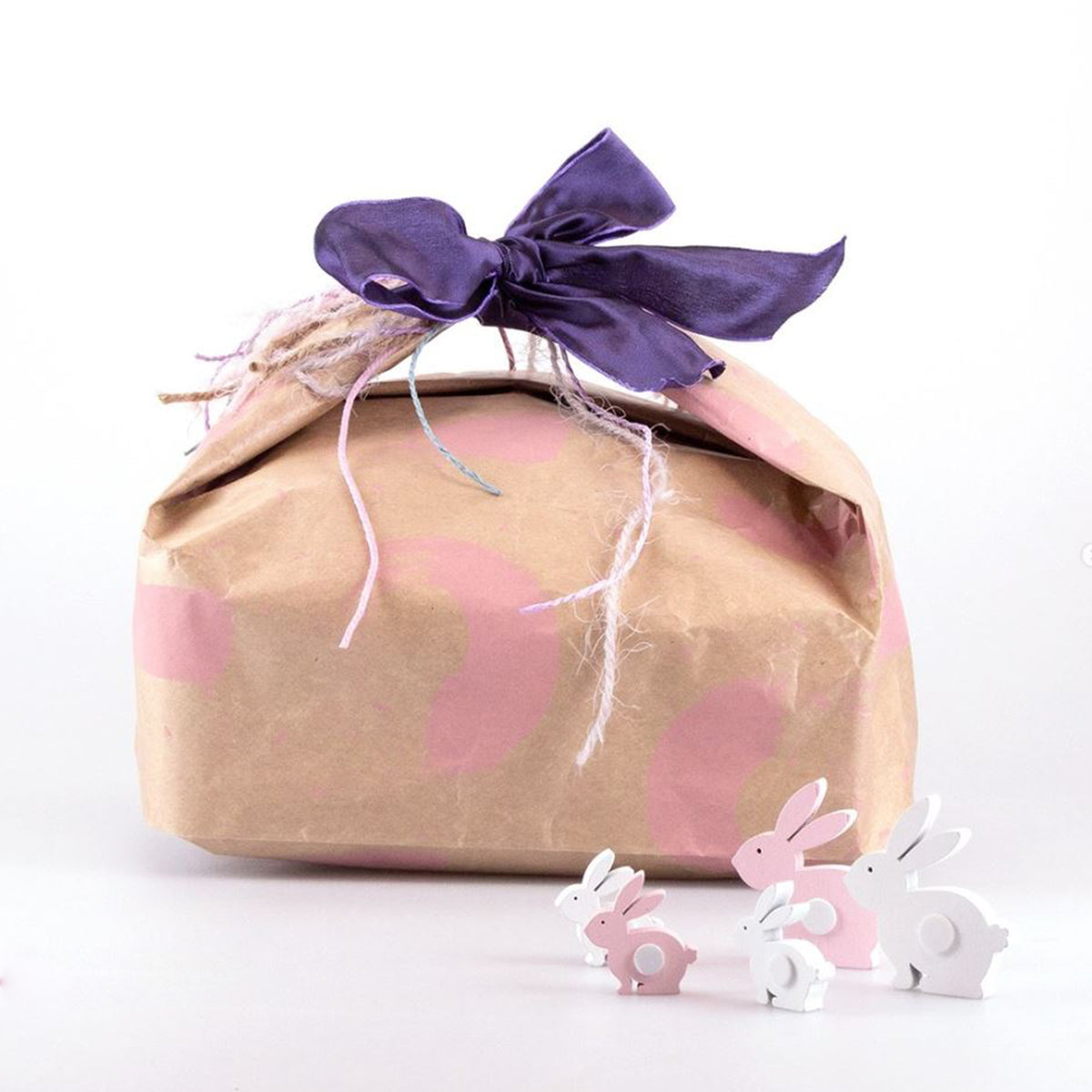 Sacchetto strip porta colomba color avana con fantasia rosa, fiocco viola, accanto a coniglietti di legno 