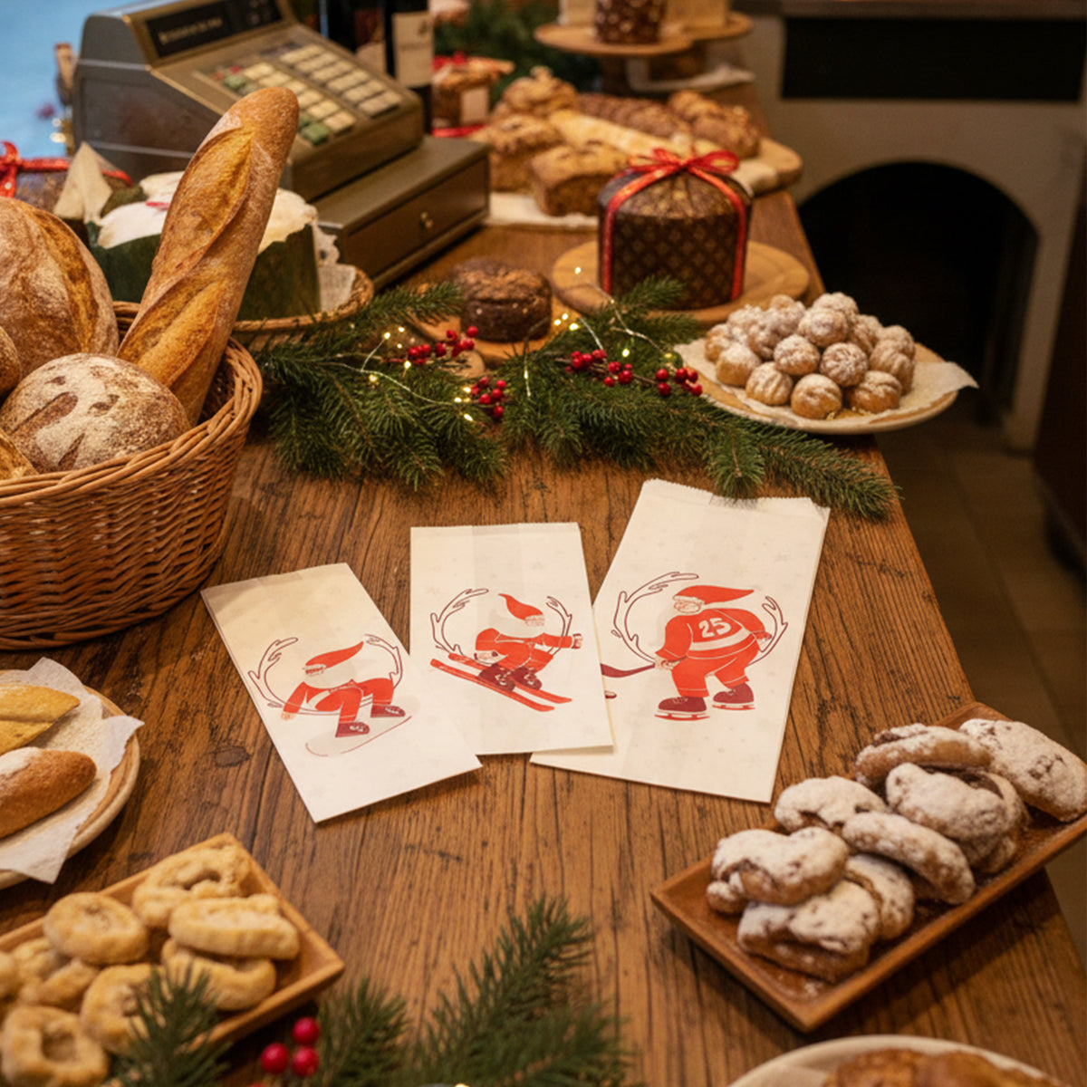 Tre sacchetti di carta per il pane, con disegnato babbo natale, su un tavolino tra pani, dolci in una ambientazione natalizia 