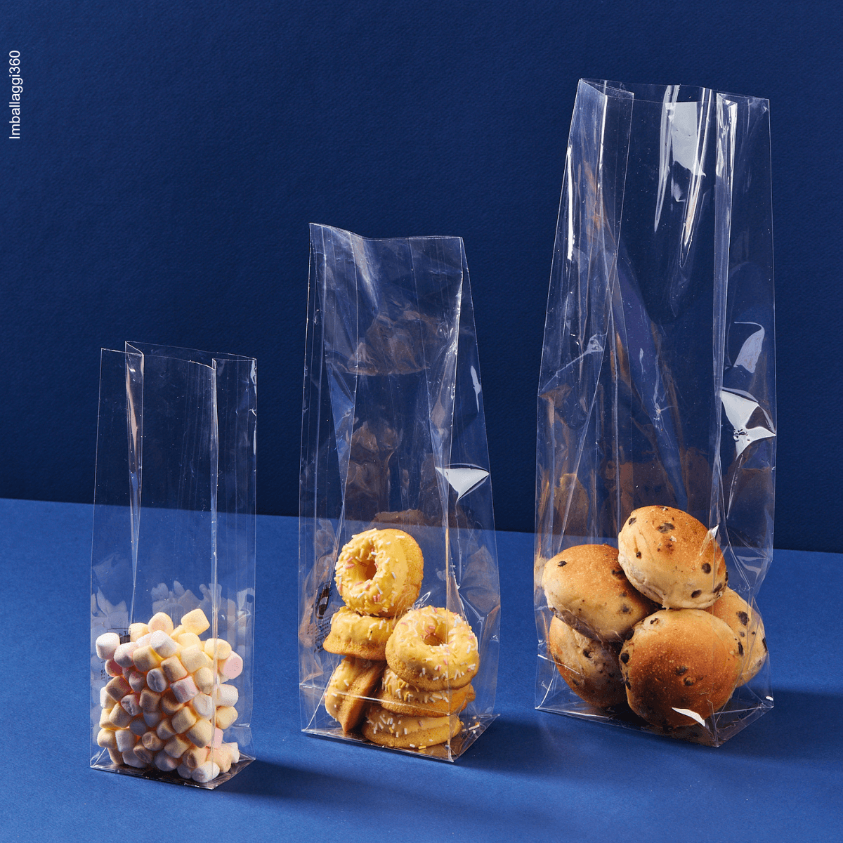 Sacchetto a fondo quadro, trasparente, ripieno di caramelle e dolci, aperti, varie misure. Sfondo blu 