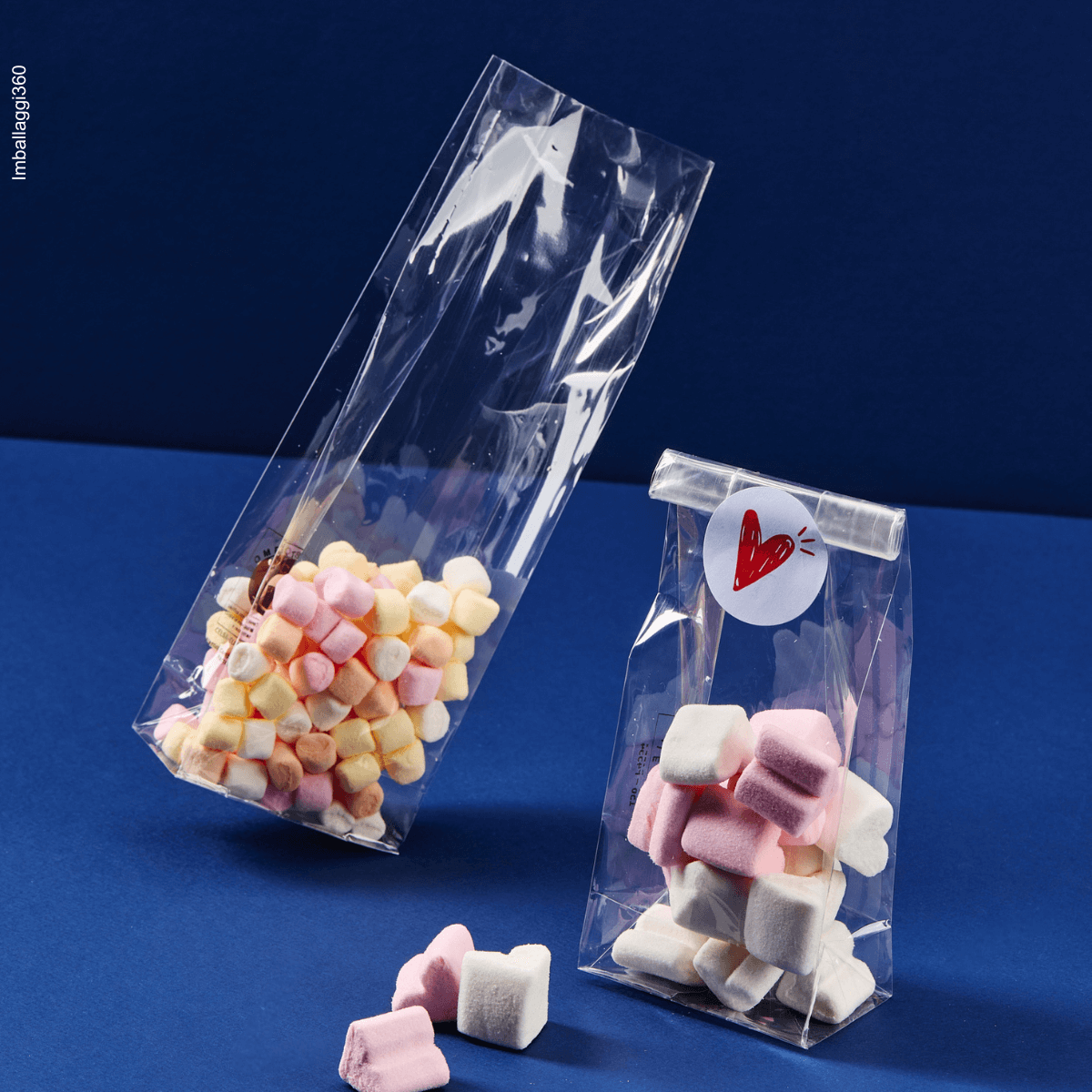 Sacchetto a fondo quadro, trasparente, ripieno di caramelle. Mostrato aperto e chiuso su sfondo blu 