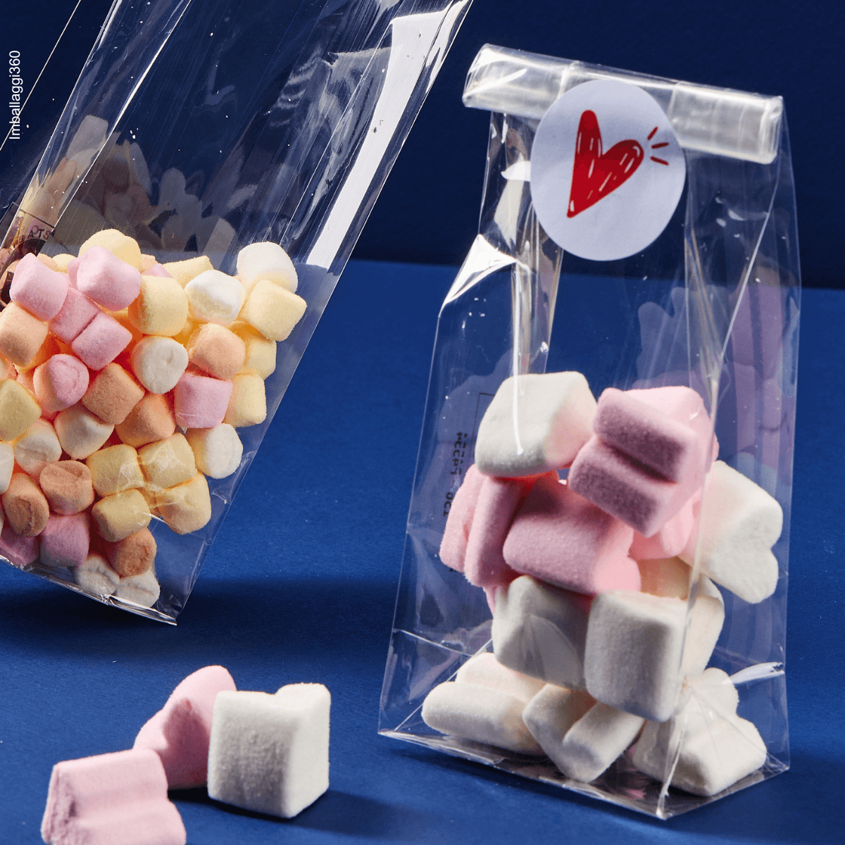 Sacchetto trasparente, fondo quadro, ripieno di dolci, dettaglio di uno aperto e centralità di uno chiuso con etichetta cuore . Sfondo blu
