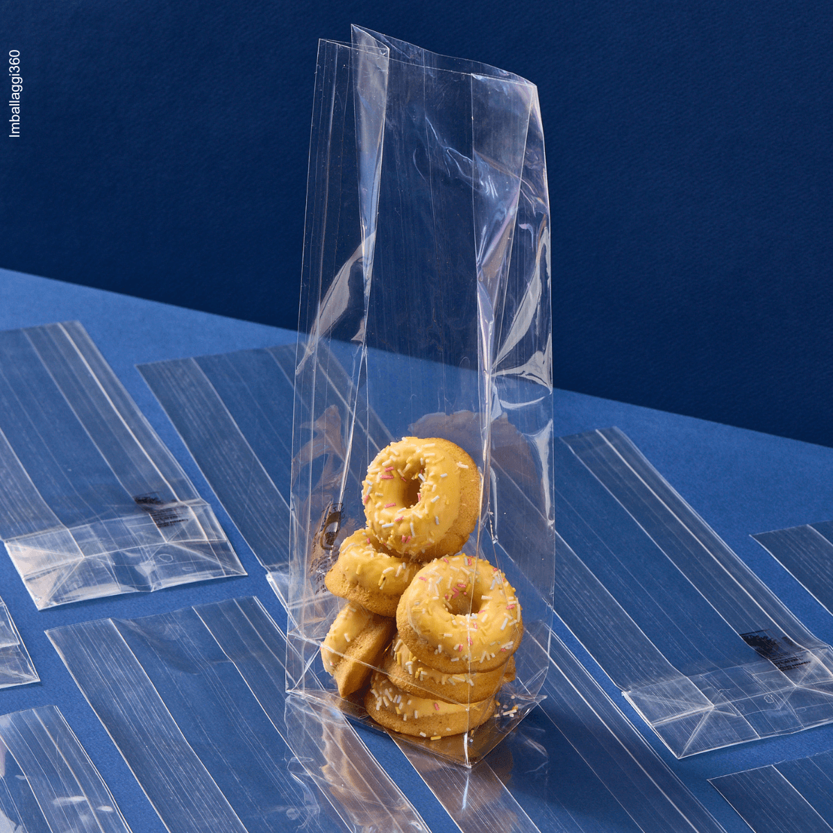 Sacchetto a fondo quadro, trasparente, ripieno di dolci, aperto, su sacchetti chiusi e stesi. Sfondo blu 