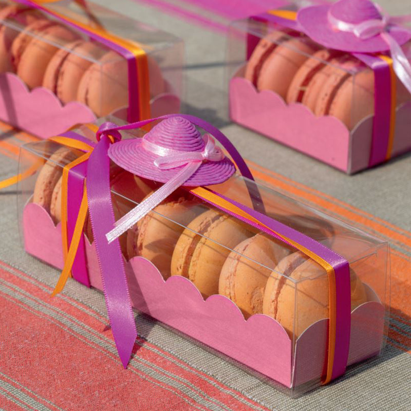 Scatola per macarons,  fucsia e coperchio trasparente  ripiena di macarons con fiocco viola, su un tavolo assieme ad altre 
