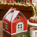 Scatola per panettone a forma di casetta rossa su tavolo di legno accanto a elementi natalizi e lucine