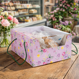 Scatola porta colomba con finestra con tema floreale su base rosa, con dentro una colomba su un tavolo di legno di una pasticceria 
