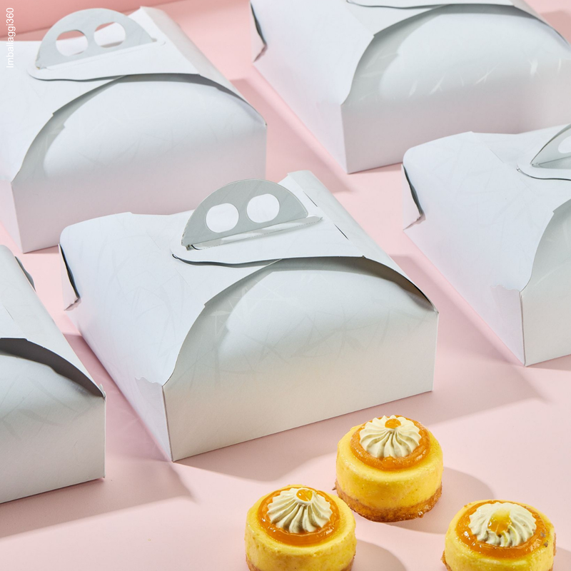 Scatola porta torte bianca, chiusa assieme ad altre tutte disposte in fila e sempre chiuse. Tre pasticcini di fronte 