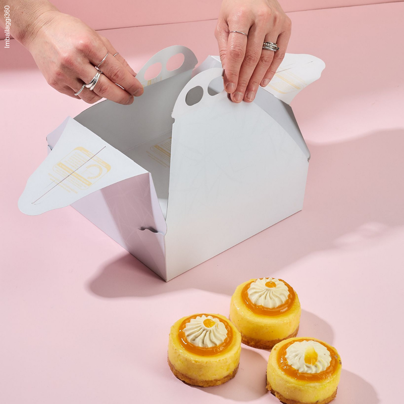 Scatola porta torte bianca nell'atto di essere chiusa da due mani e 3 pasticcini di fronte 
