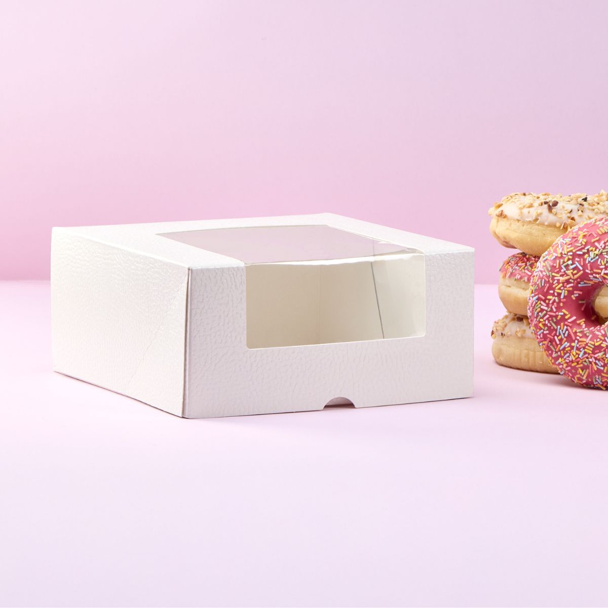 Scatola porta torte bianca, con finestra, vuota, con ciambelle impilate vicino su sfondo rosa