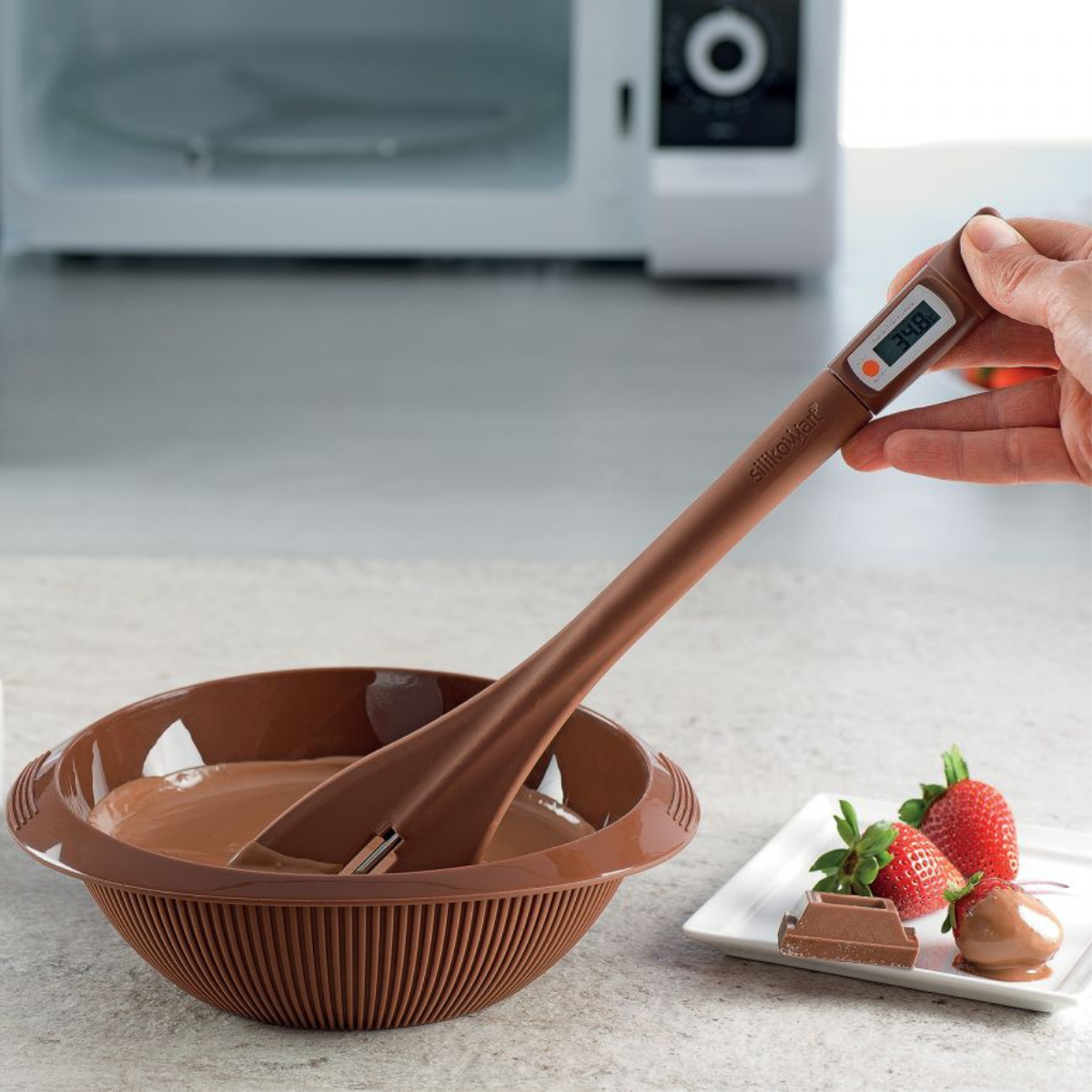 Mano che regge un termometro per cioccolato dentro una vaschetta in cui sta sciogliendo cioccolato 