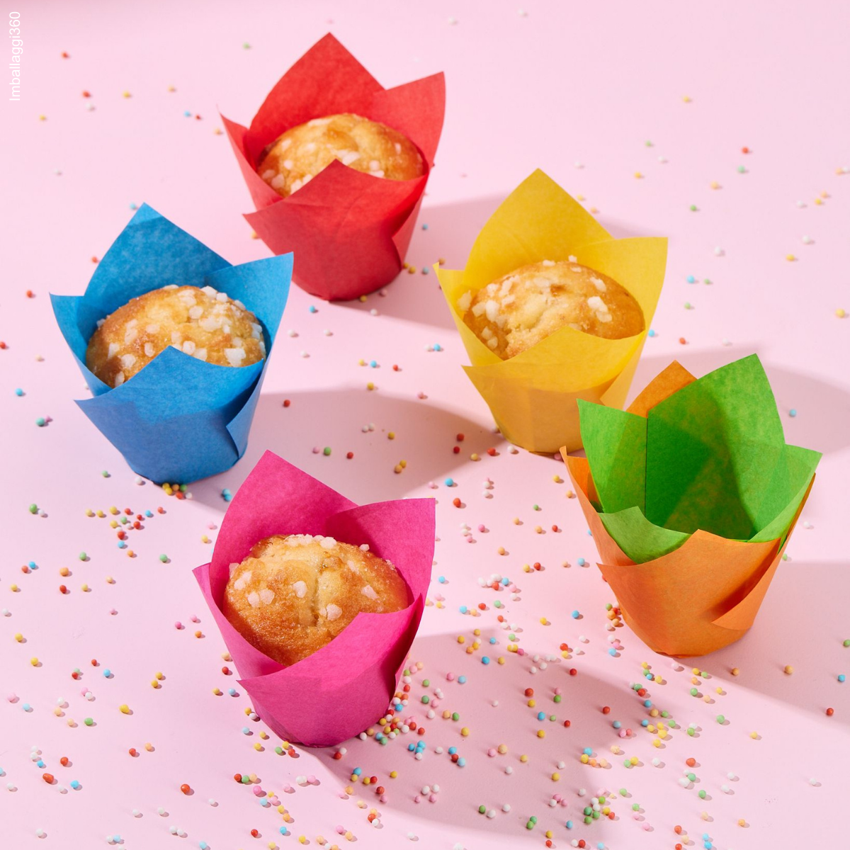 Pirottini colorati, alcuni pieni di dolci e due impilati. Tutti su tavola rosa con zuccherini colorati sparsi 