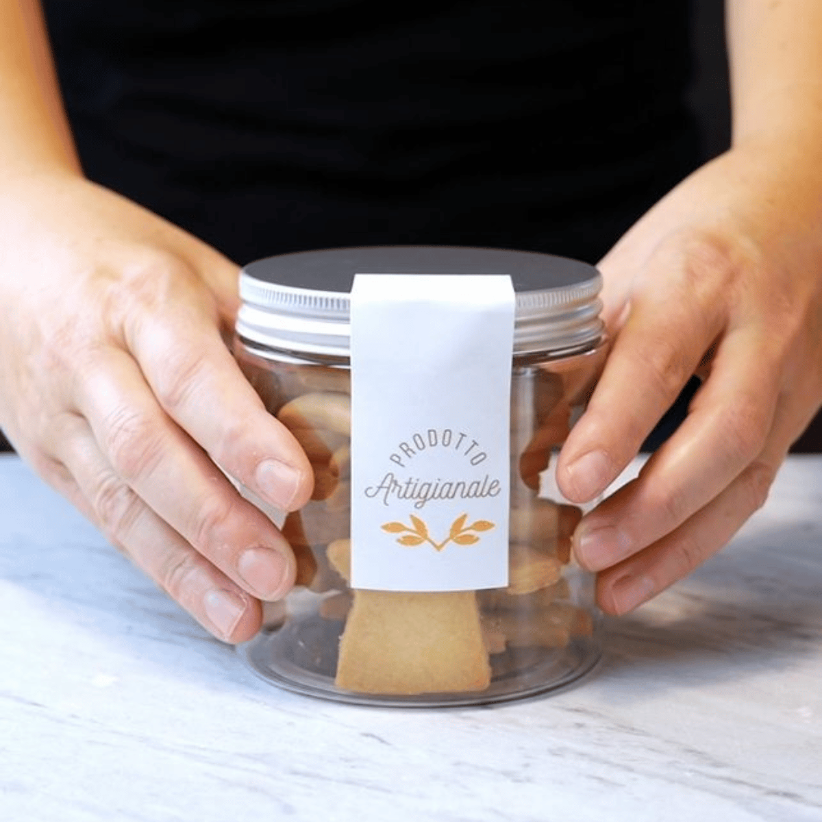 Vasetto trasparente con tappo in alluminio pieno di biscotti ed adesivo prodotto artigianale retto tra due mani 