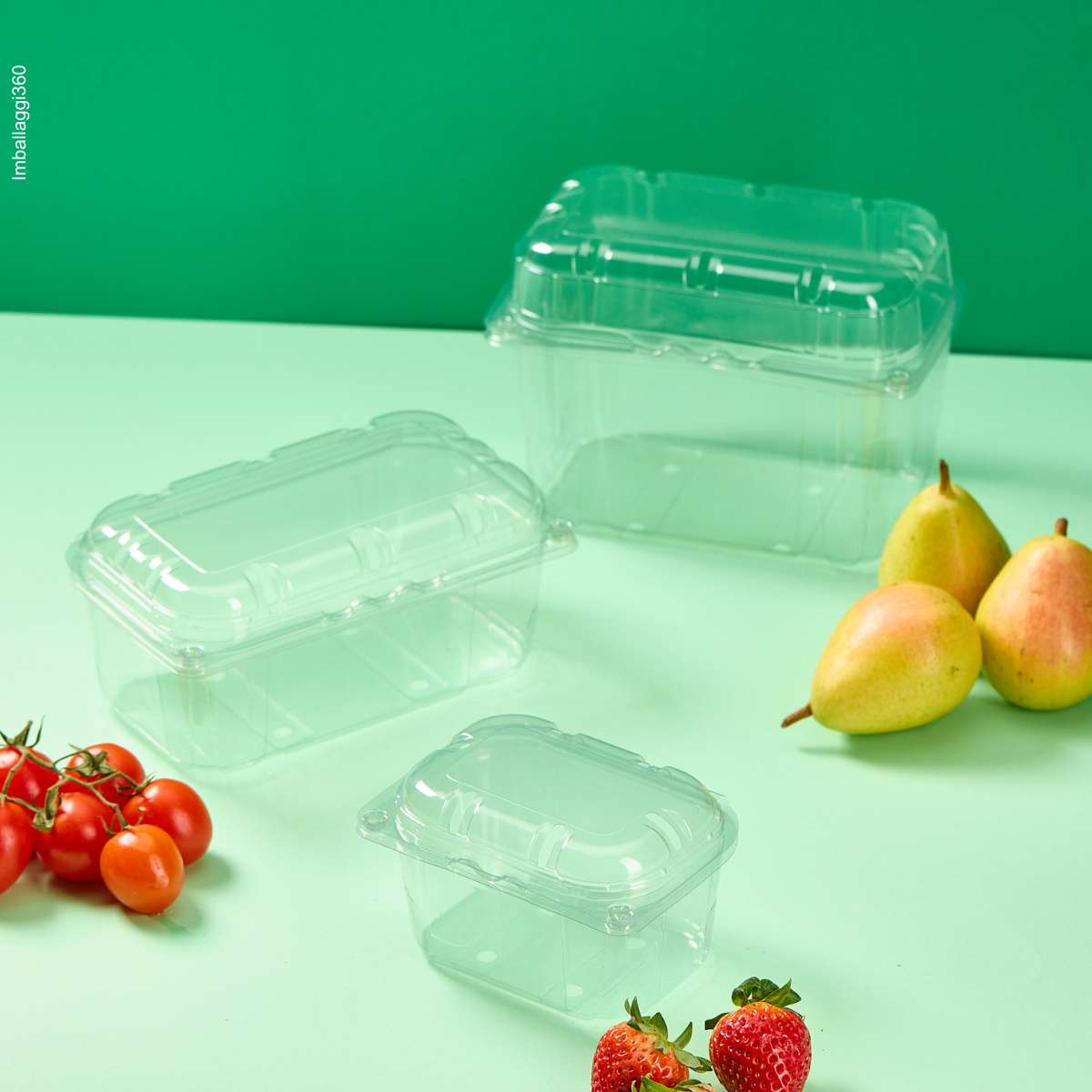 Vaschetta trasparente per frutta e verdura, varie misure e vuote, su sfondo verde, con accanto pere, fragole e pomodorini