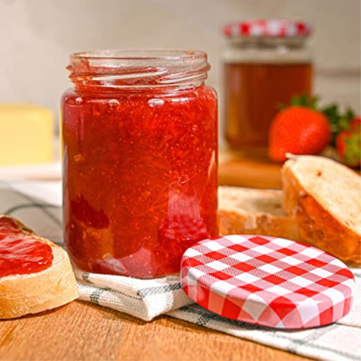 vasetto di vetro con dentro confettura, tappo a scacchi appoggiato vicino, su un tavolo con fette di pane e frutta 