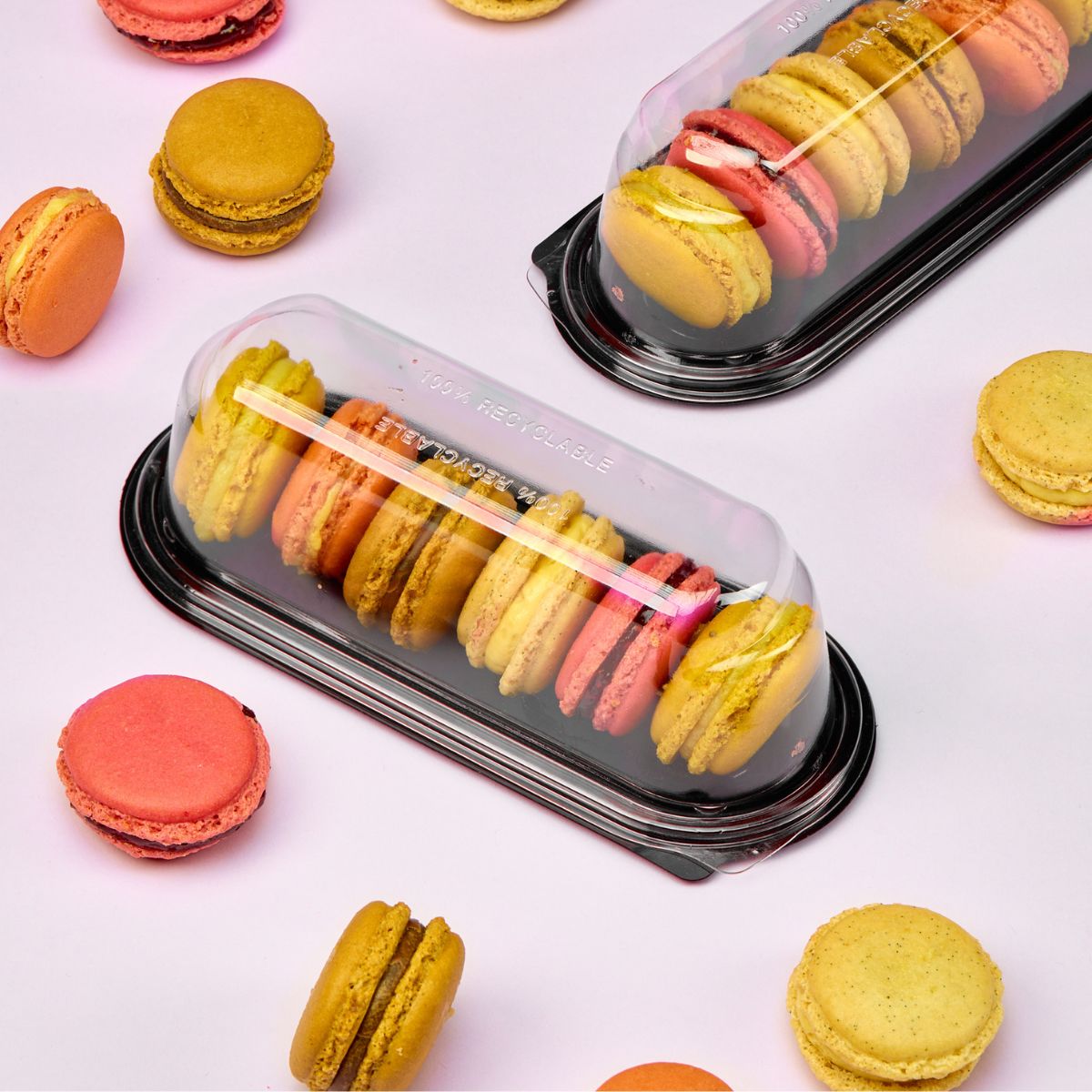 Vaschetta per monoporzioni, base nera coperchio trasparente, piena di macarons, e macarons sparsi su sfondo rosa