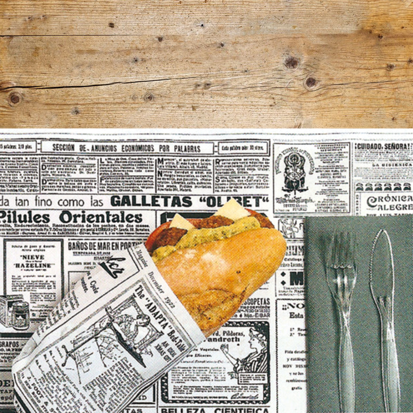 Dettaglio di tavola con tovaglietta americana bianca con scritte nere, panino avvolto dalla stessa carta e tovagliolo di carta con due posate di plastica trasparente 