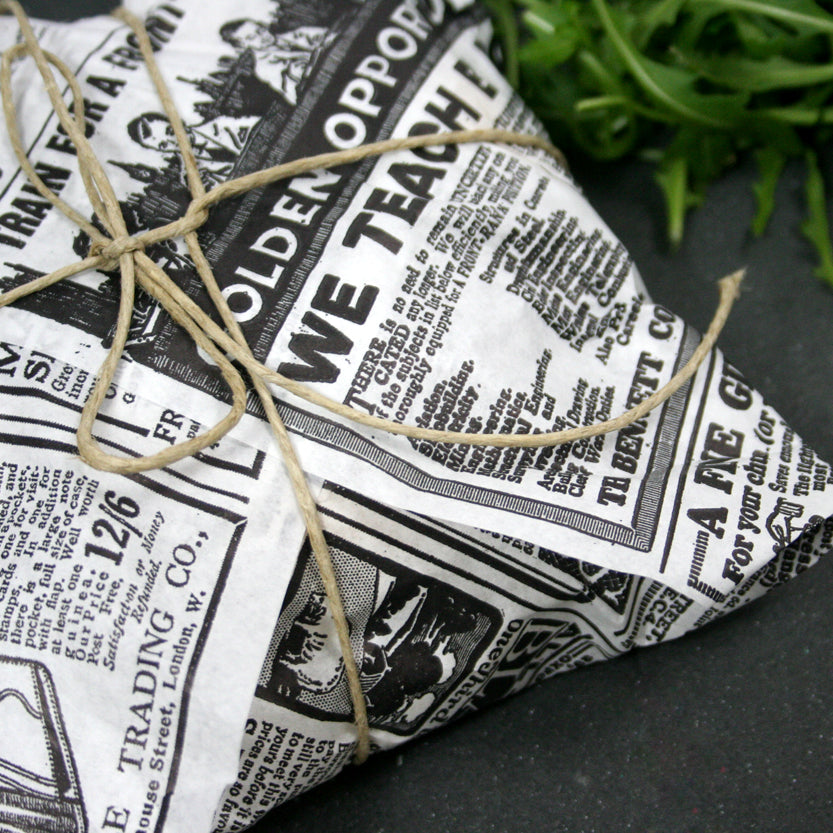 Dettaglio pacchettino chiuso con carta alimentare bianca con scritte nere e spago su tavolo nero vicino a della rucola 