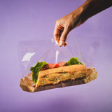 Mano che tiene sacchetto di plastica trasparente  fondo in cartone avana con dentro un panino 