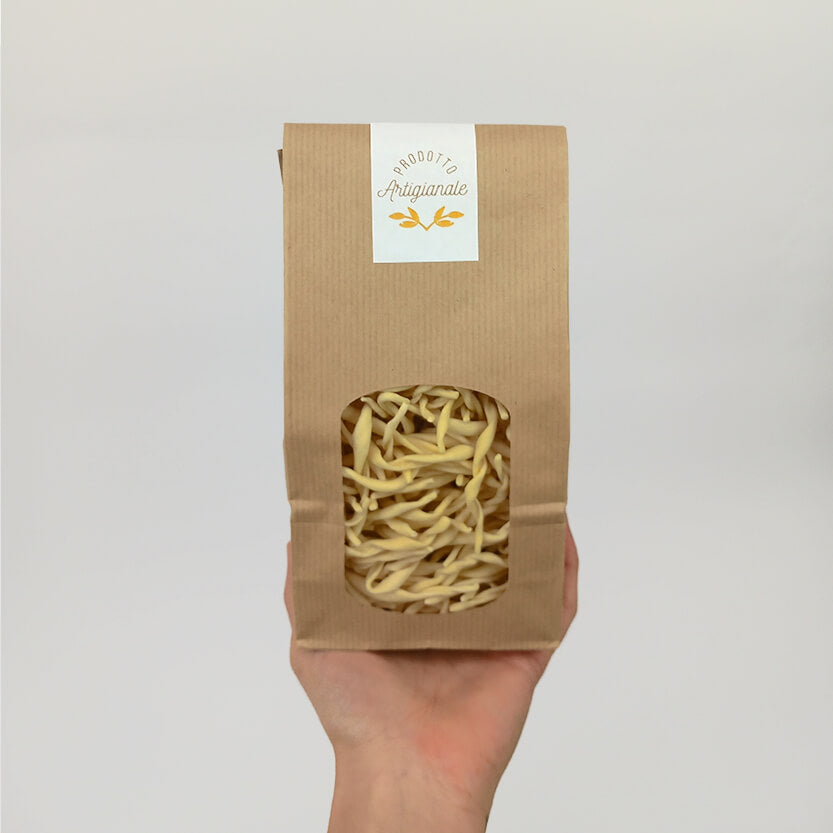 Mano che regge sacchetto di carta color avana con finestra pieno di strozzapreti con etichetta rettangolare bianca Prodotto artigianale 