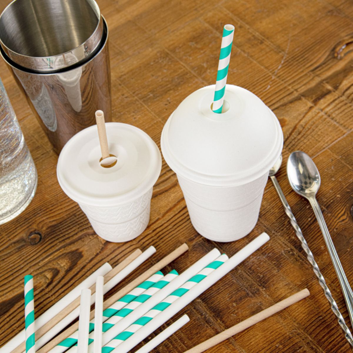 Bicchieri da sporto, due misure diverse, con coperchio a cupola uno l'altro piatto, con foro per cannucce, su tavola di legno, con cannucce ed elementi di mixology 