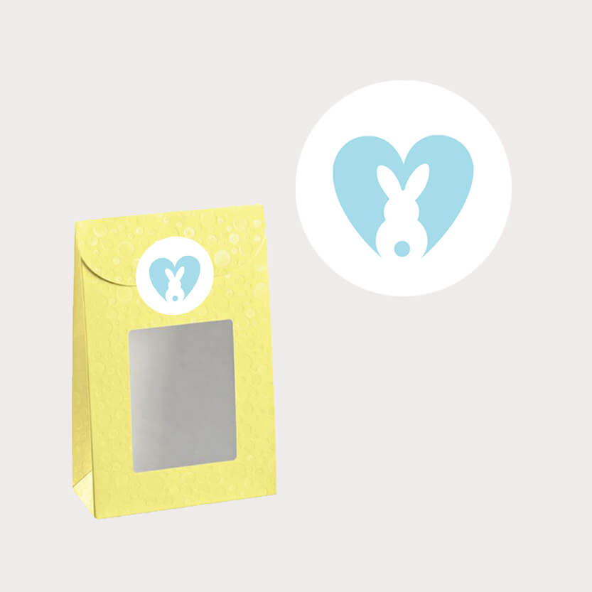 Etichetta bianca tonda con coniglietto stilizzato dentro cuore azzurro mostrata singola e su una confezione verde con finestra 