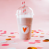Bicchiere di plastica con cupola con frappe e due cannucce a righe bianche e rosse con una etichetta tonda con un cuore tra coriandoli