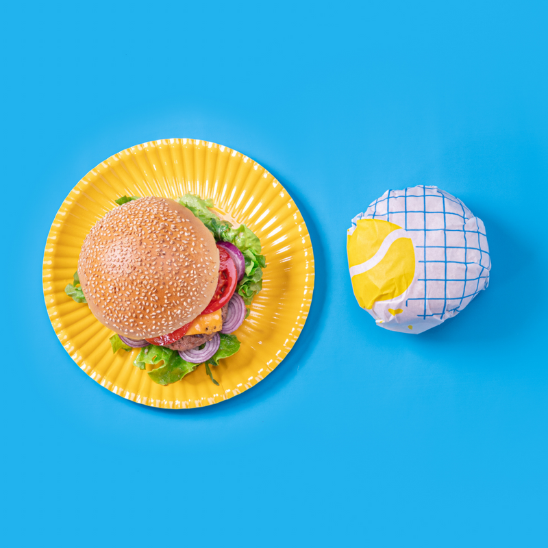 Piatto monouso di carta giallo con sopra panino e di fianco un panino avvolto in carta antigrasso reticolata blu , tutto visto dall'alto