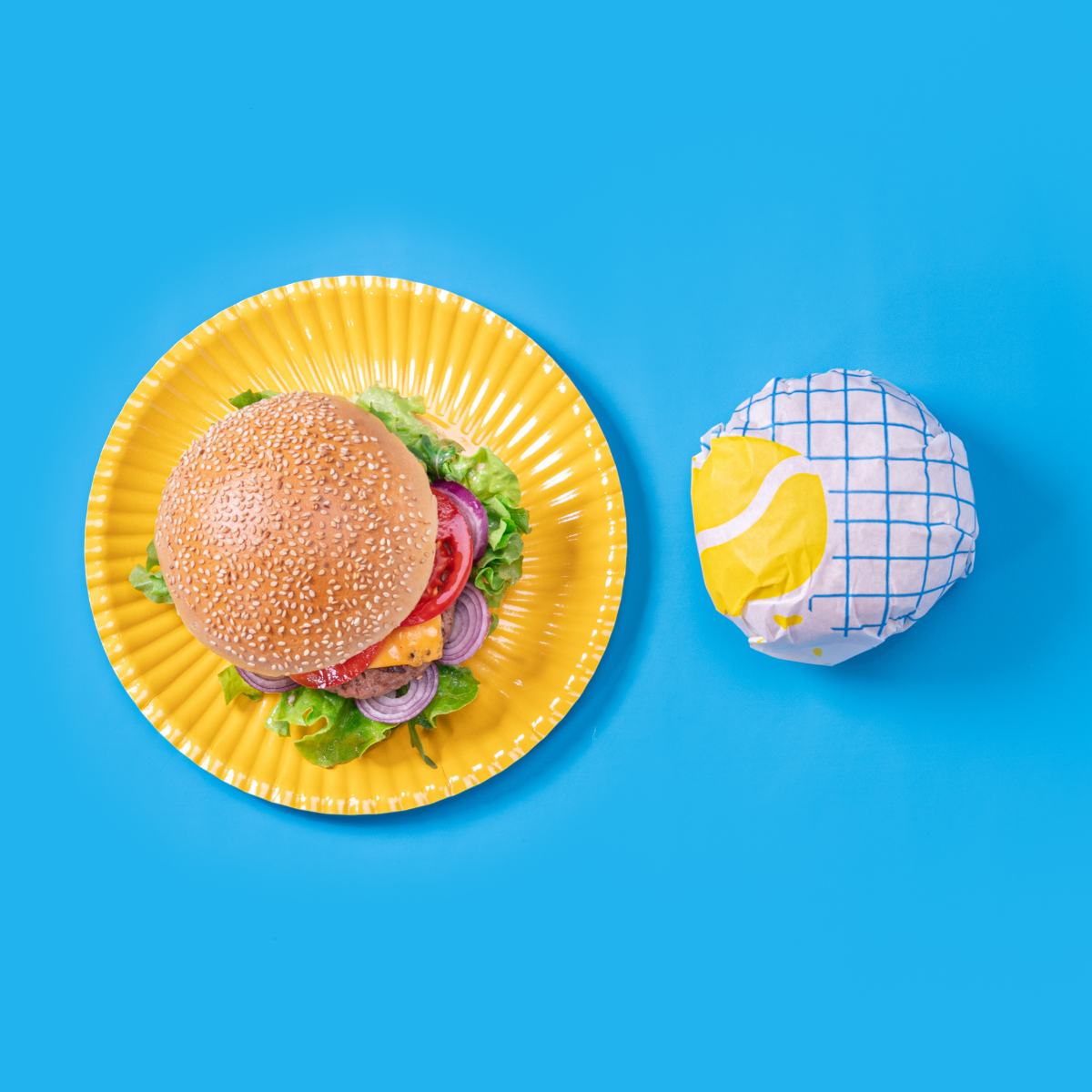Piatto monouso di carta giallo con sopra panino e di fianco un panino avvolto in carta antigrasso reticolata blu , tutto visto dall'alto