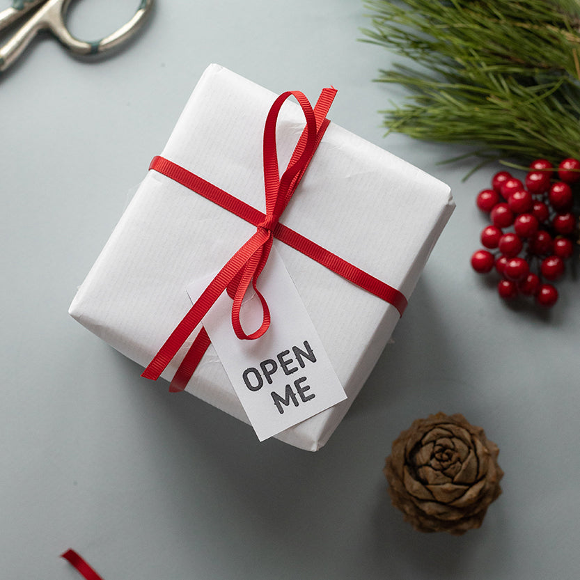 Pacco avvolto da carta bianca e fiocco rosso con cartellino open me 