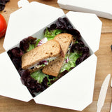 Scatola di cartone per cibo da asporto mostrata aperta con insalata e panini dentro, su un tavolo di legno 