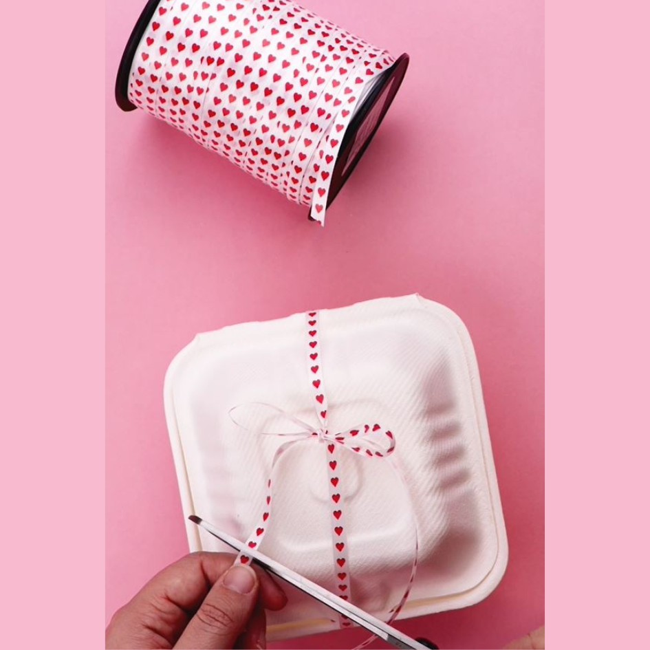 Bobina di nastro bianco con cuoricini rossi e mano che usa il nastro per chiudere una scatola porta panino bianca 