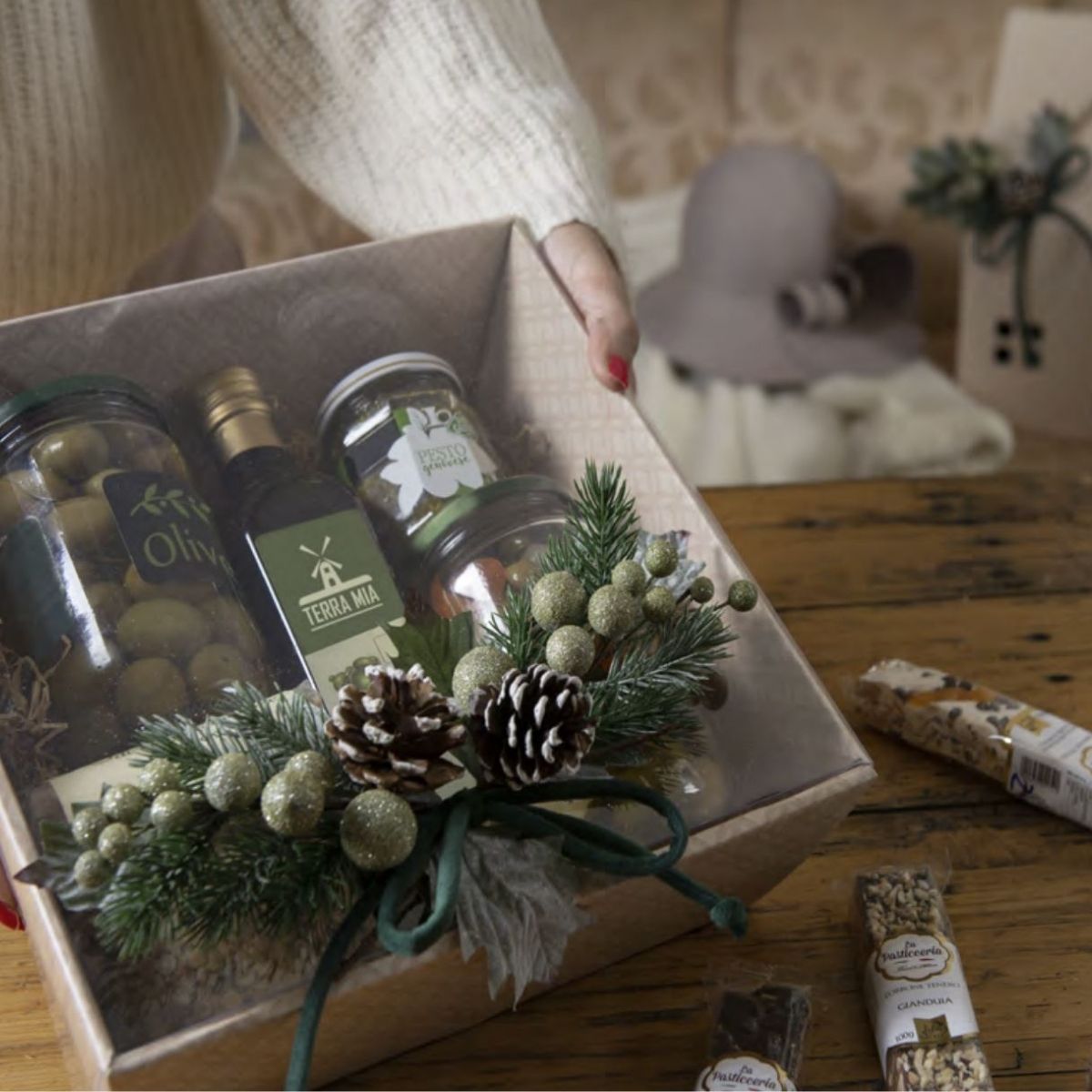 Mani che reggono su un tavolo una scatola quadrata con coperchio trasparente piena di vasetti e addobbata in maniera natalizia